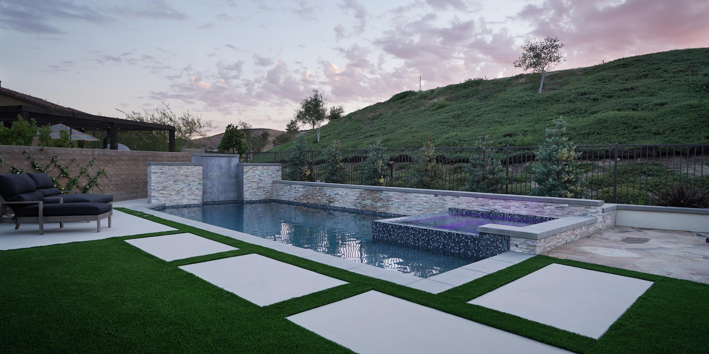 A serene backyard scene with a modern pool and spa area. The pool is bordered by stone tiles and features a waterfall. Two dark lounge chairs sit on the left, on a patio with artificial grass accents. In the background, a green hillside under a pastel evening sky completes the peaceful atmosphere.