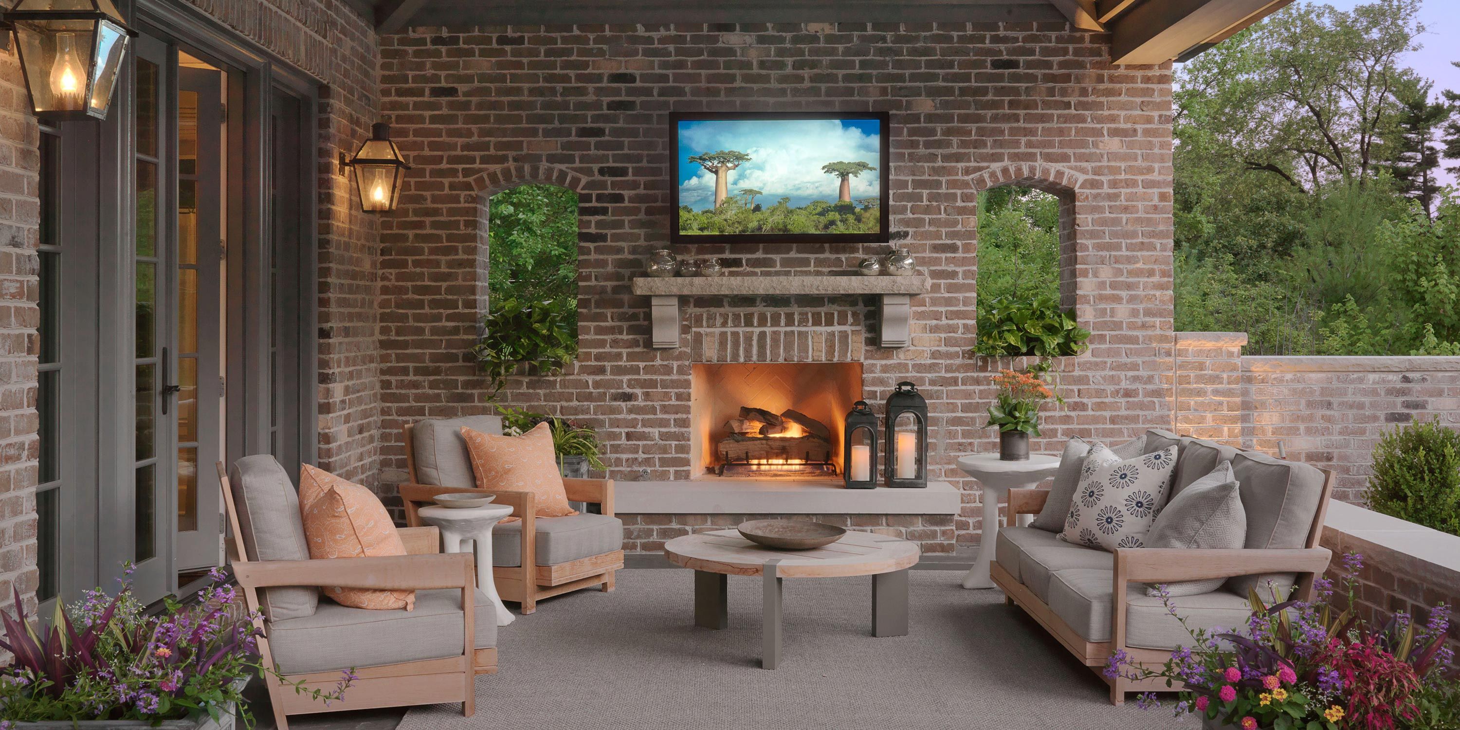 A cozy outdoor patio with a brick fireplace, seating area, a Seura outdoor TV,and lush greenery.