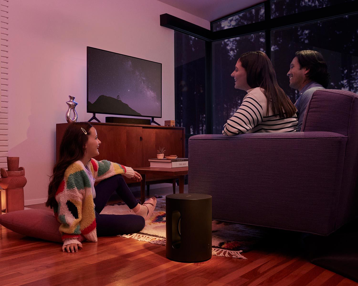 A family watching TV together in a cozy living room at night, with a young girl sitting on the floor and a Sonos speaker visible near the TV cabinet.