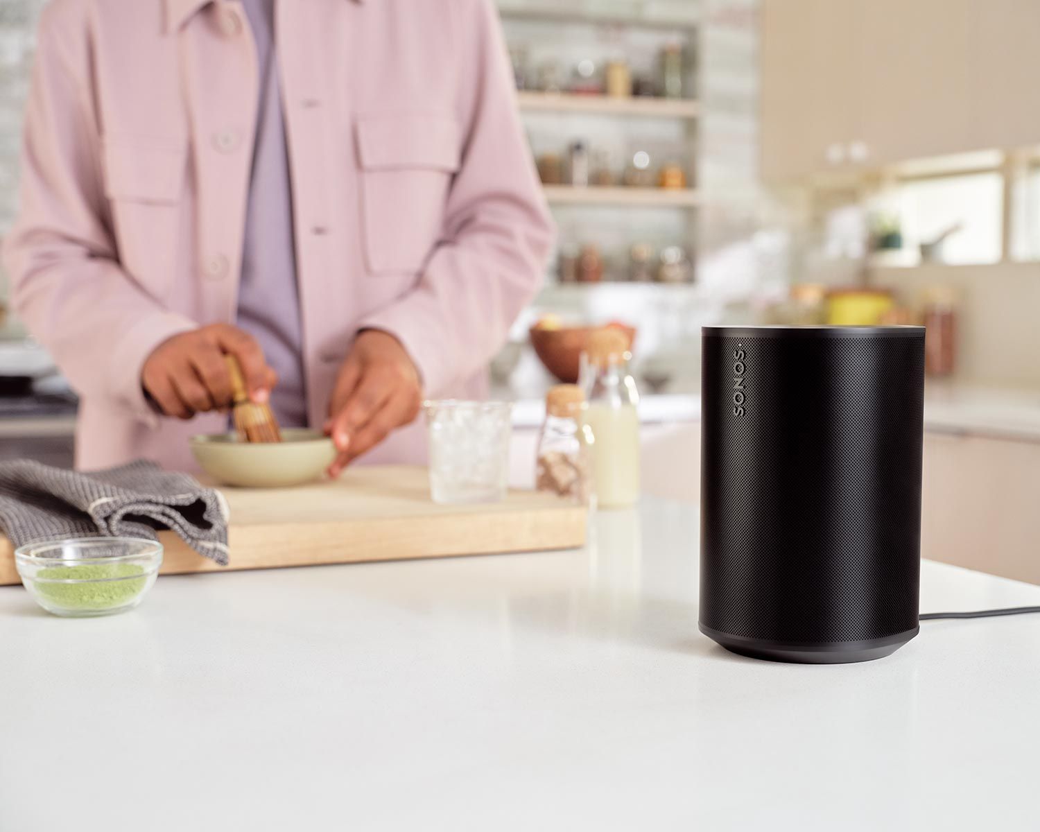 A close-up of a Sonos speaker on a kitchen counter, with a person preparing food in the background.