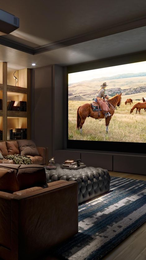 A cozy Sony home theater featuring a large screen showing an image of a cowboy on horseback in an open field. The room is equipped with comfortable seating, a tufted ottoman, and bookshelves with ambient lighting.