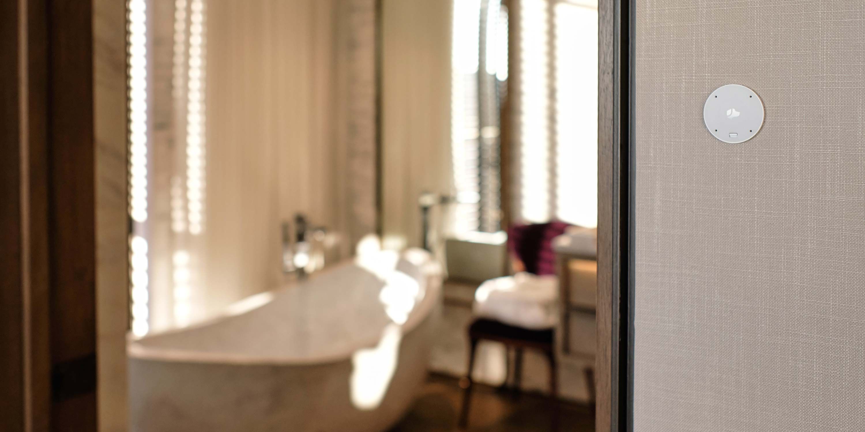 A Josh AI wall-mounted circular device with a dog logo, installed on a textured wall in a modern, well-lit bathroom with a blurred background of a bathtub and elegant furnishings.