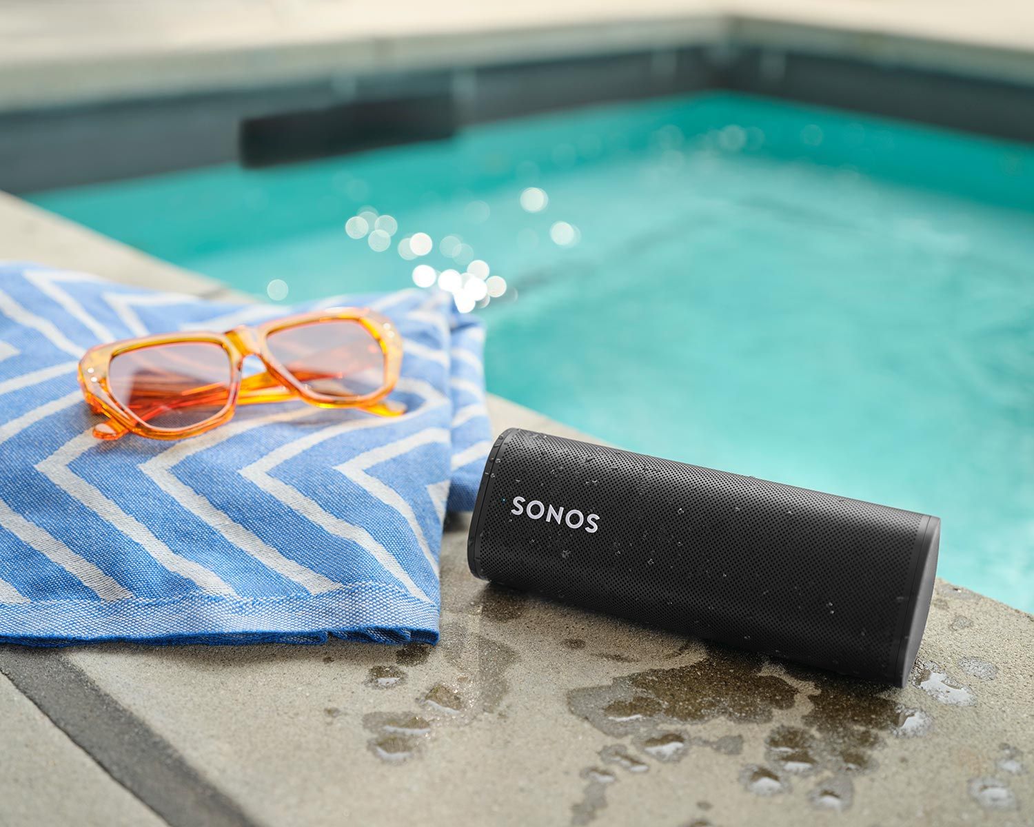 A Sonos portable speaker placed next to a swimming pool with a pair of sunglasses on a towel nearby.