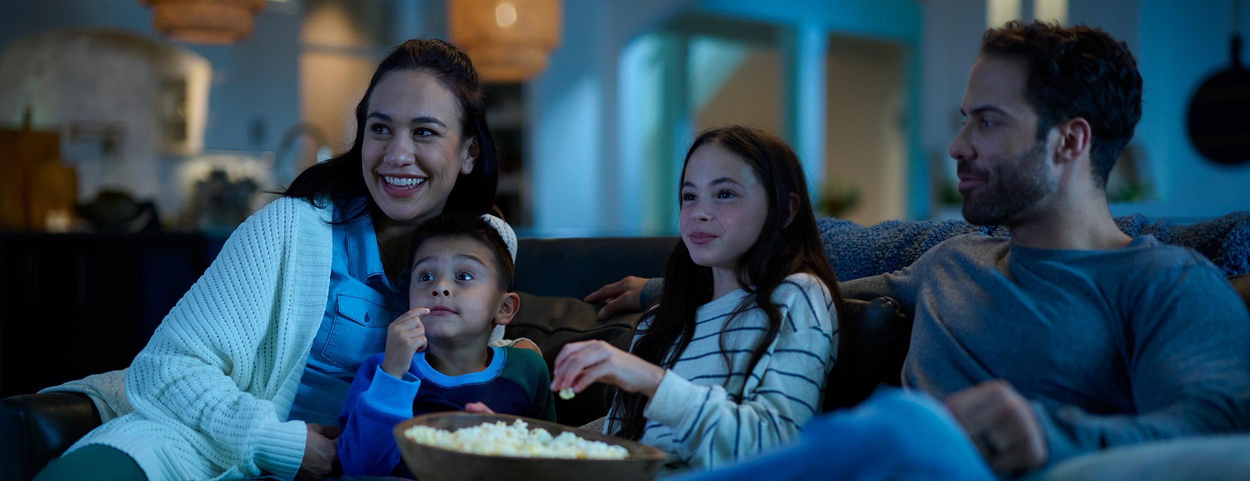 A family of four sits together on a cozy couch, smiling and watching a movie while sharing a bowl of popcorn in a warmly lit living room.