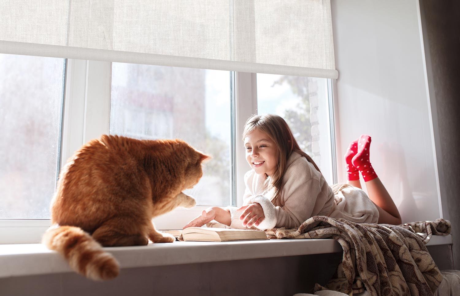 A young girl lies on a windowsill, smiling and interacting with a large ginger cat. Sunlight streams through the window, highlighting the cozy, bright scene. The window is partially covered by a light-colored Crestron shade.