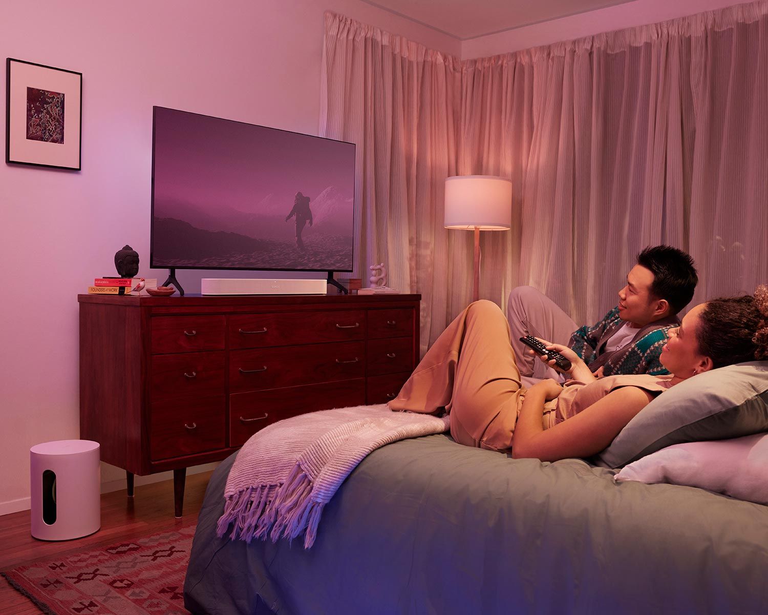 A couple relaxing in bed while watching TV in a softly lit bedroom with Sonos Mini and Arc.