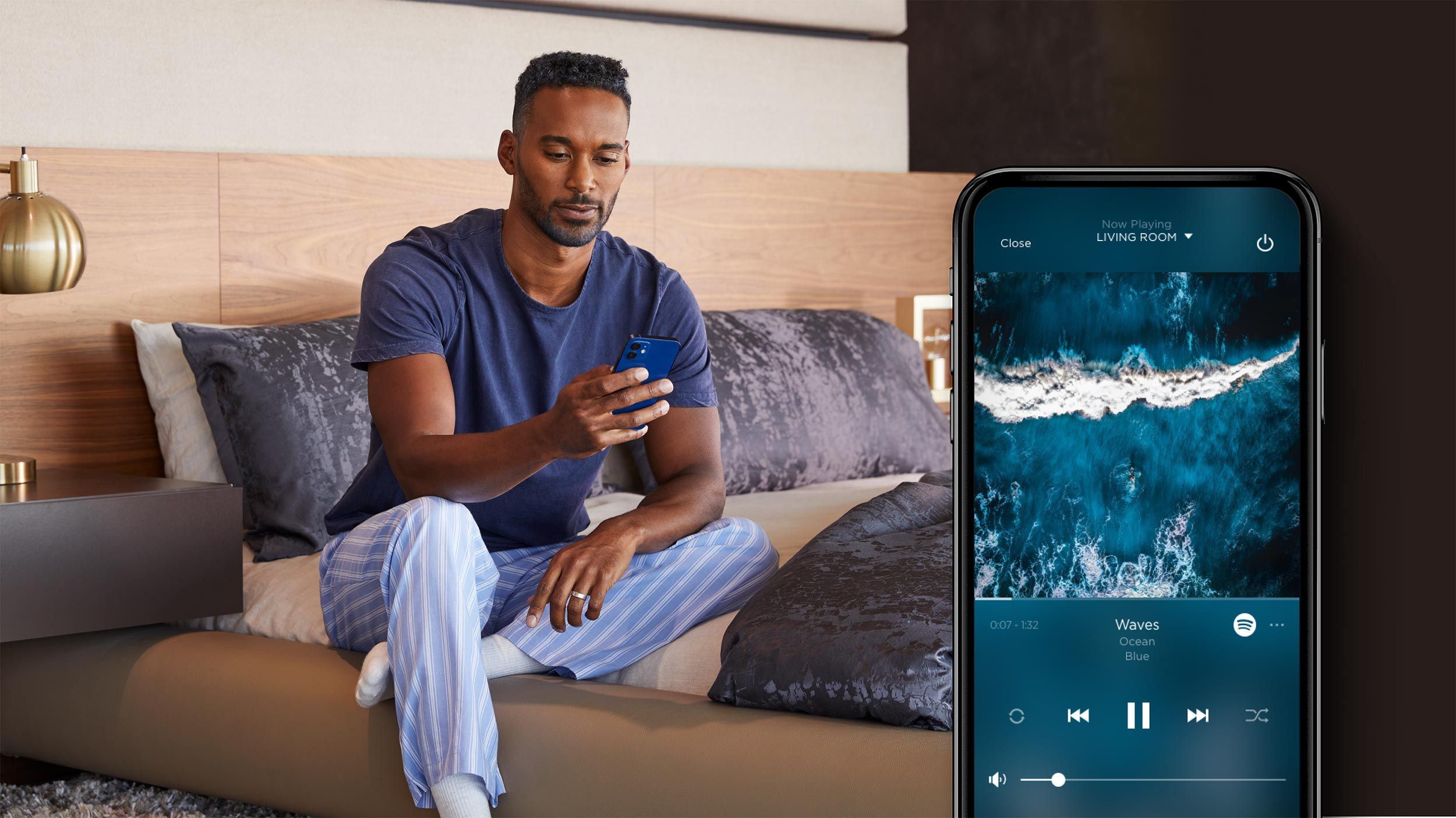 A man in comfortable pajamas sits on a bed, using a smartphone to control music, with the phone screen displaying a song titled "Waves" and an image of ocean waves.